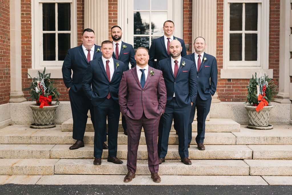 Seven men in suits stand on stone steps outside a brick building. The man in front wears a burgundy checked suit, while the others wear navy suits. Two large planters with holiday decorations flank the entrance.
