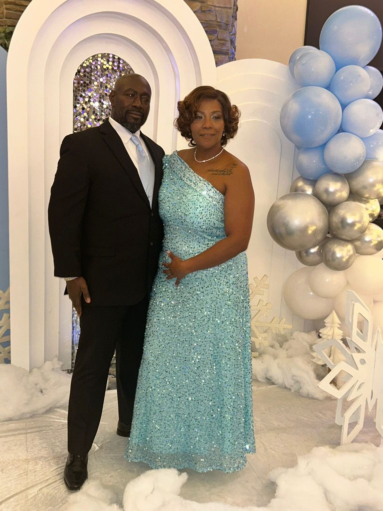 A man in a black suit and a woman in a sparkly blue one-shoulder gown stand together in front of an arch with silver and blue balloons, snowflake decorations, and artificial snow.