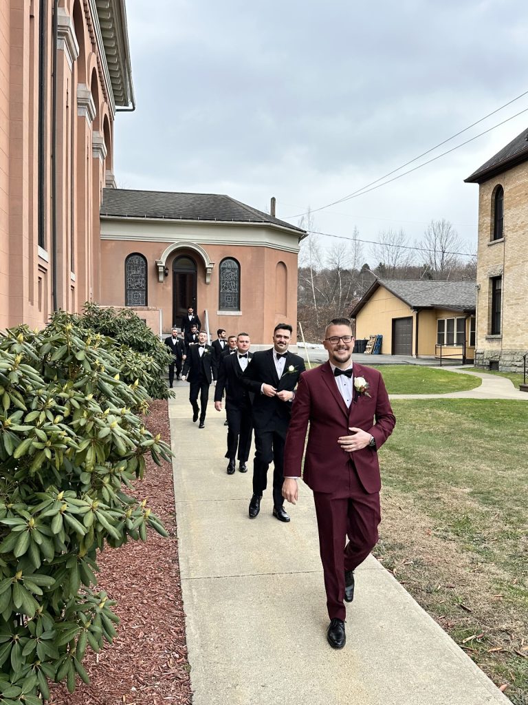 A group of men in tuxedos, led by a man in a burgundy suit holding a bouquet, walk outside a church on a cloudy day, smiling as they head down a sidewalk.