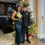 A young woman in a green dress and a young man in a black suit with a green shirt stand in front of a decorated Christmas tree, smiling, holding a bouquet, in a cozy living room.