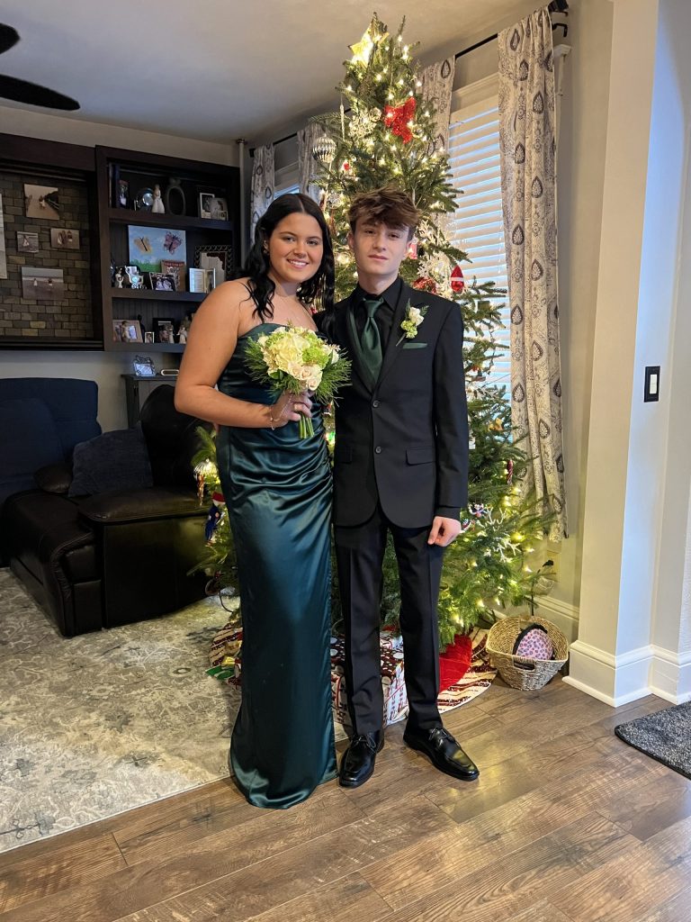 A young woman in a green dress and a young man in a black suit with a green shirt stand in front of a decorated Christmas tree, smiling, holding a bouquet, in a cozy living room.