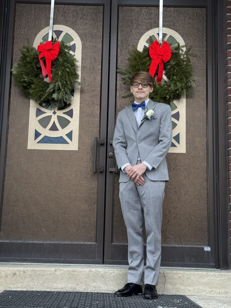 A young person in a light gray suit, blue bow tie, and glasses stands in front of double doors decorated with holiday wreaths featuring red bows.