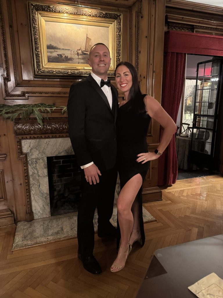 A man in a black tuxedo and a woman in a sleeveless black dress with a thigh-high slit pose together, smiling, in front of a fireplace and wooden paneled wall with a sailboat painting above the mantle.