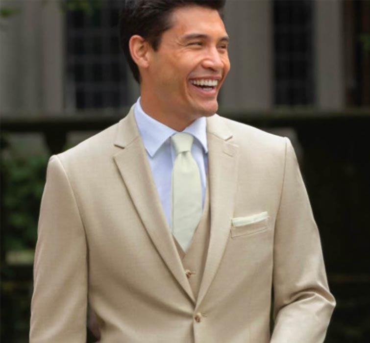 A man in a light beige suit, white shirt, and pale tie stands outdoors, smiling happily. He has dark hair and is wearing a pocket square. The background is blurred, with hints of greenery and a building.