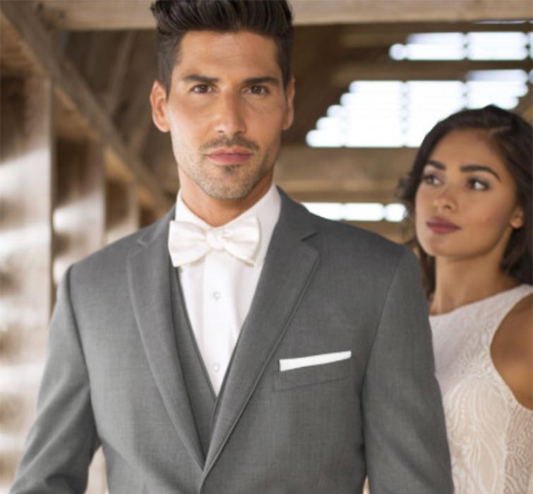 A man in a grey suit with a white bow tie stands in the foreground, while a woman in a white dress stands slightly behind him in a softly lit, rustic setting.