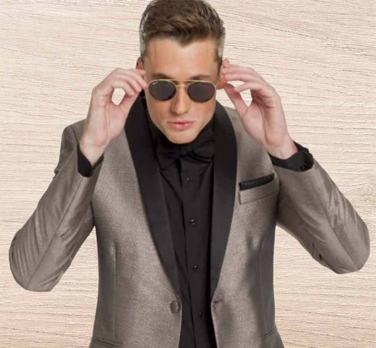 A man wearing a shiny gray suit with black lapels and a black shirt adjusts his sunglasses while facing the camera, standing in front of a light wood-textured background.