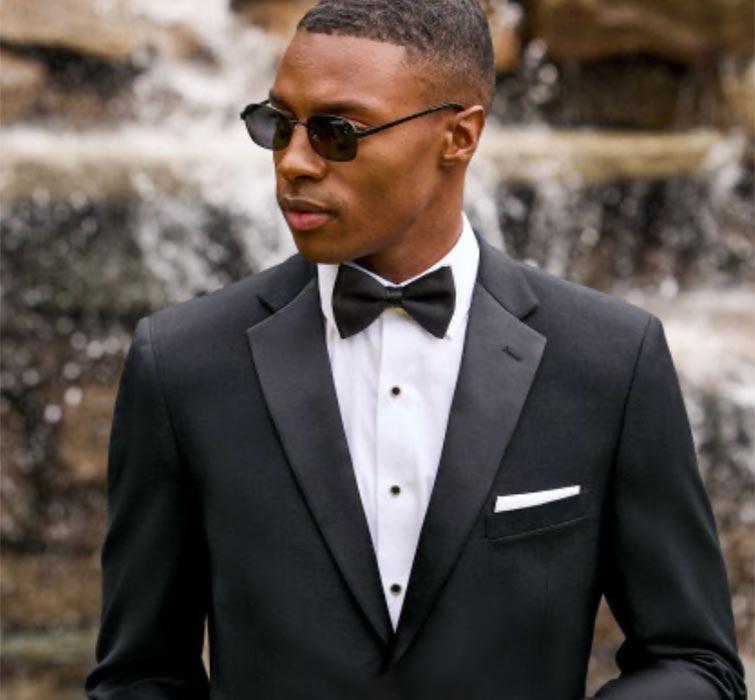 A man wearing sunglasses, a black tuxedo, white dress shirt, and black bow tie stands in front of a blurred waterfall, looking to the side.