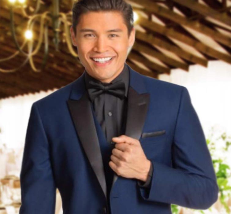 A smiling man in a blue tuxedo with black satin lapels and a black bow tie stands indoors under wooden rafters with soft lighting.