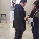 A man wearing a black suit is being fitted by a woman with a measuring tape in a clothing store. The man stands still while the woman adjusts his jacket. Mannequins and sale signs are visible in the background.