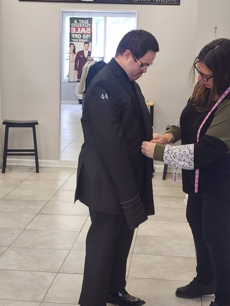 A man wearing a black suit is being fitted by a woman with a measuring tape in a clothing store. The man stands still while the woman adjusts his jacket. Mannequins and sale signs are visible in the background.