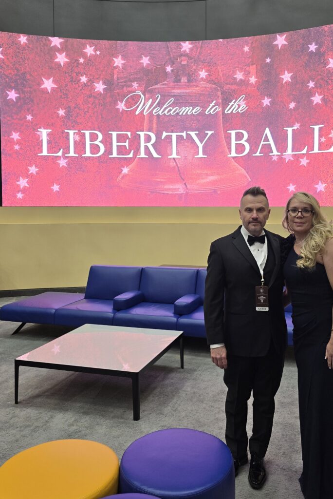 A man in a tuxedo and a woman in a black dress pose together in front of a large screen that reads Welcome to the Liberty Ball, with a starry red background. Modern blue and yellow furniture is in the foreground.