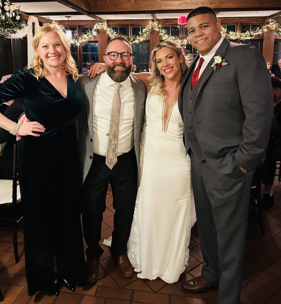 Four people pose closely together and smile at a wedding. One woman wears a black dress, a man wears glasses and a gray suit, the bride wears a white gown, and the groom wears a gray suit with a red tie. Festive lights decorate the background.