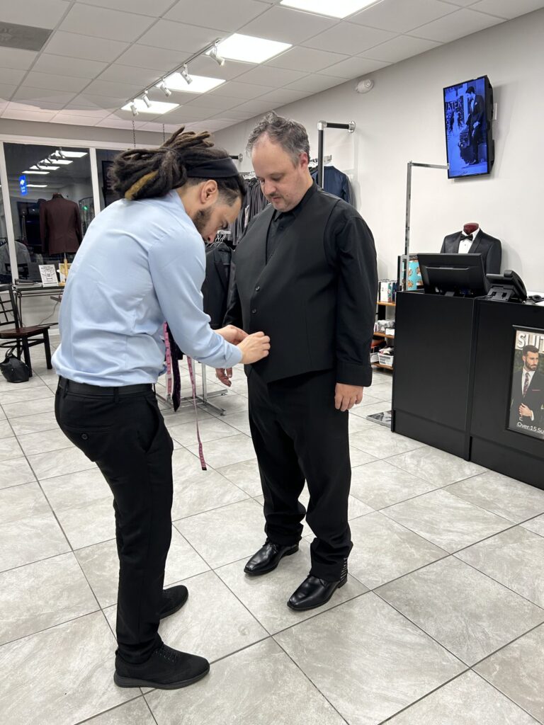 A man in a light blue shirt adjusts the vest of another man dressed in black formalwear inside a modern clothing store with suits and tuxedos on display.