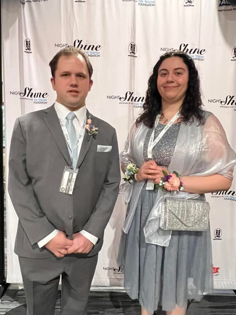 A man in a gray suit and a woman in a silver-blue dress stand together, smiling, in front of a “Night to Shine” event backdrop. Both wear event badges and corsages.