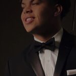A young man dressed in a black tuxedo with a black bow tie and a white pocket square, smiling and looking to the side in a dimly lit setting.