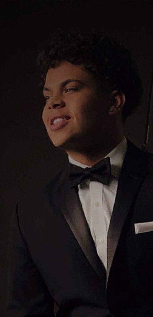 A young man dressed in a black tuxedo with a black bow tie and a white pocket square, smiling and looking to the side in a dimly lit setting.