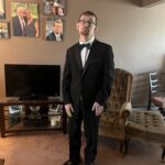 A man wearing a black tuxedo with a bow tie stands in a living room, holding a small green object. Behind him are family photos on the wall, a TV on a stand, and an upholstered chair beside him.