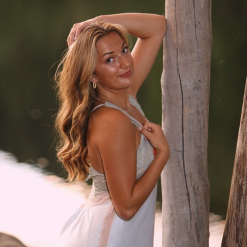 A woman with wavy blonde hair in a light dress poses outdoors by a wooden post, with her arm raised and a gentle smile, set against a blurred natural background.