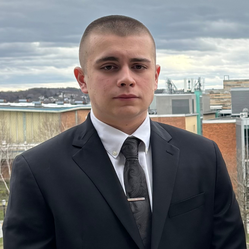 A young man with closely cropped hair wears a black suit, paisley tie, and white shirt, standing outside with an overcast sky and modern buildings in the background. He has a neutral expression.