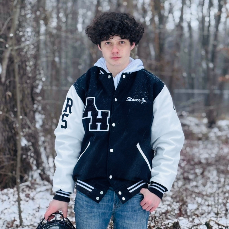 A teenage boy with curly hair stands in a snowy forest wearing a black and white varsity jacket and jeans, holding a sports helmet in his left hand. Snow lightly covers the ground and trees in the background.