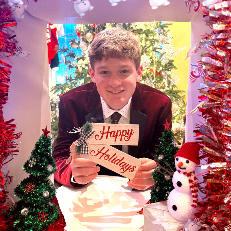 A young man in a red velvet jacket and tie smiles, holding a Happy Holidays sign. He is framed by festive decorations, including tinsel, small Christmas trees, and a snowman, with a decorated tree in the background.