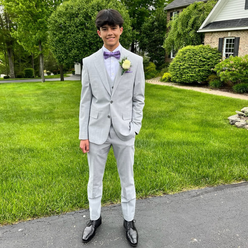 A young man stands on a driveway wearing a light gray suit, lavender bow tie, and black dress shoes, with a white boutonnière. He is smiling, with green grass and houses in the background.