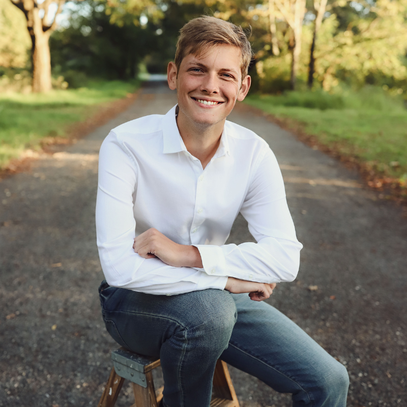 A young man in a white shirt and jeans sits on a wooden stool in the middle of a tree-lined path, smiling at the camera on a sunny day.