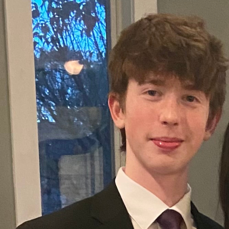 A young person with fair skin and brown hair, wearing a black suit, white shirt, and purple tie, smiles and sticks their tongue out slightly. They are standing indoors near a window with trees visible outside.