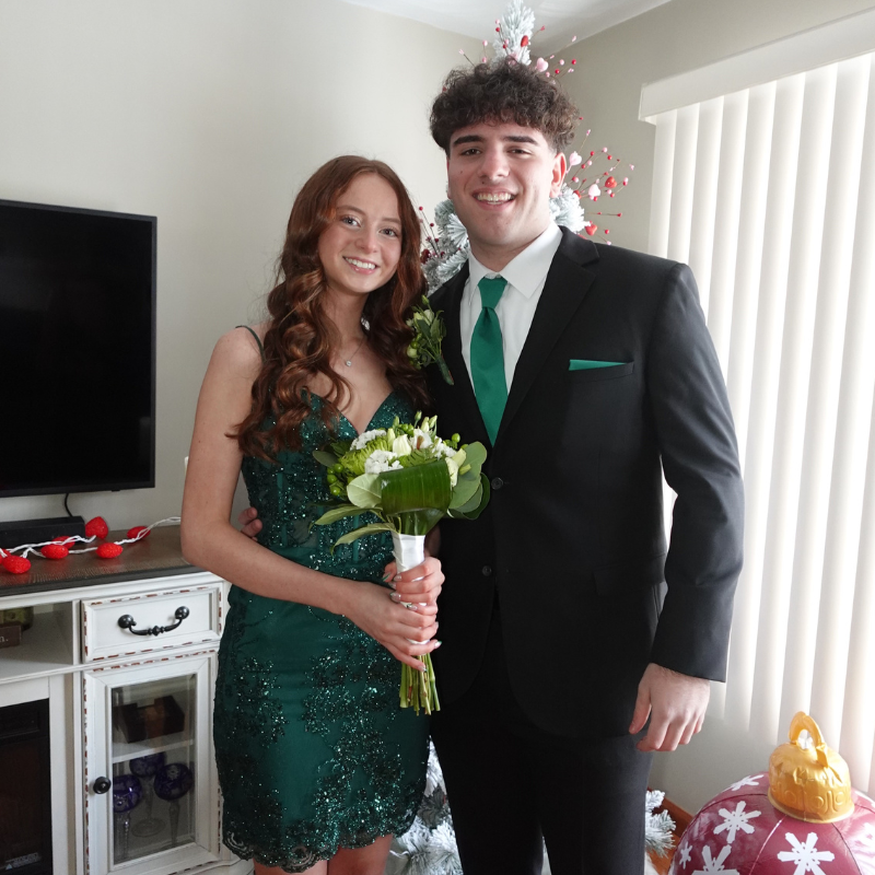 A young couple dressed formally poses indoors; the woman wears a green dress and holds a bouquet, while the man wears a black suit with a green tie. They are smiling, with a decorated tree and TV in the background.