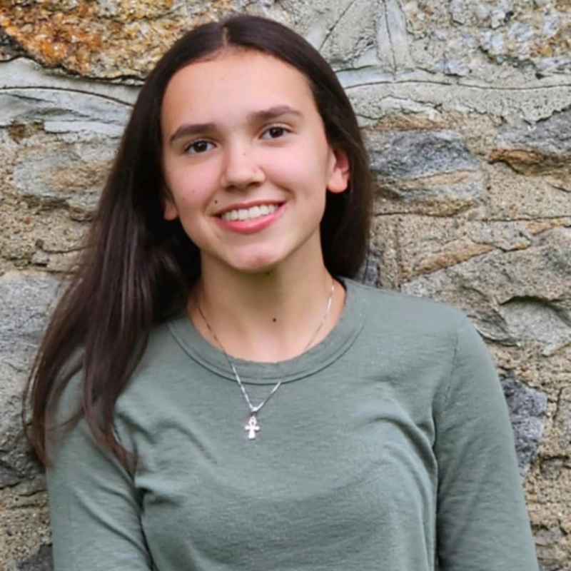 A young woman with long brown hair, wearing a green long-sleeve shirt and a silver necklace with a cross pendant, stands smiling in front of a stone wall.