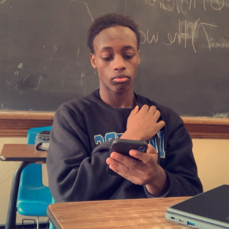 A student sits at a desk in a classroom with a chalkboard behind him, looking at his phone while resting one hand on his shoulder. A laptop is partially visible on the desk in front of him.