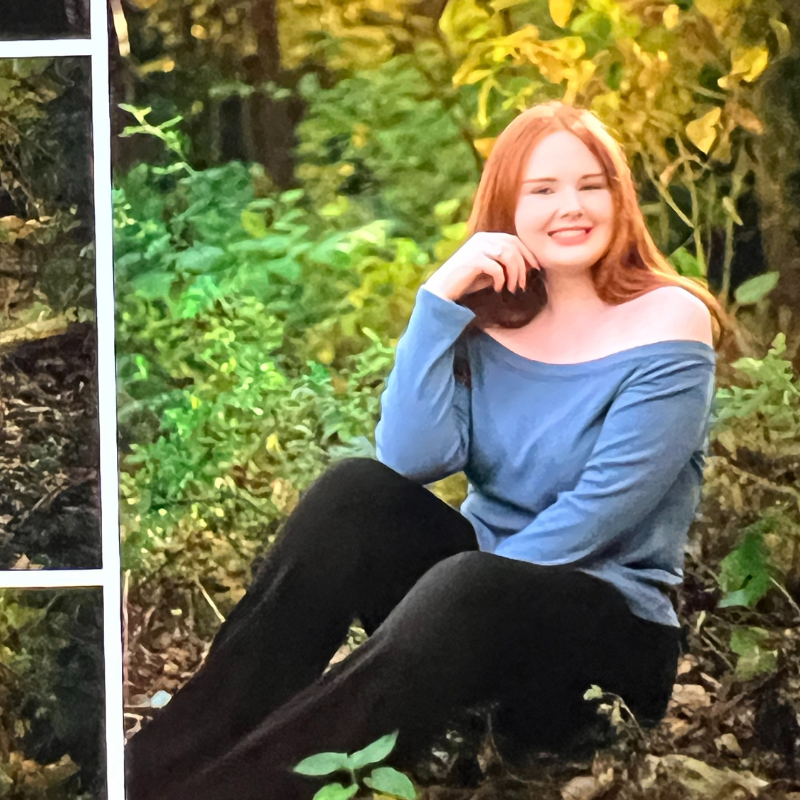 A young woman with long red hair sits outdoors on the ground, surrounded by greenery. She is smiling, wearing a blue off-the-shoulder top and black pants, with sunlight filtering through the leaves behind her.