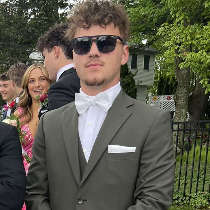 A young man wearing a gray suit, white bow tie, and sunglasses stands outside with greenery and a house in the background. Behind him, other young people are dressed formally and one girl holds a pink rose.
