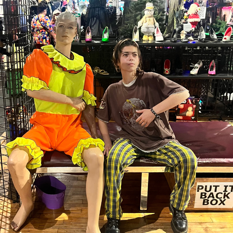 A person in a brown shirt and yellow plaid pants poses next to a mannequin wearing a neon yellow and orange ruffled outfit. They sit on a bench in front of a shoe display and a sign that reads PUT IT BACK BOX.