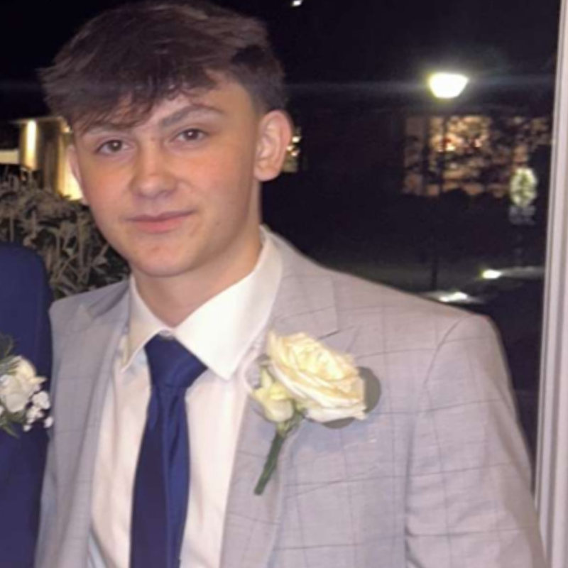 A young man wearing a light gray suit, white shirt, and dark blue tie, with a white rose boutonniere on his lapel, stands indoors at night, smiling slightly at the camera.