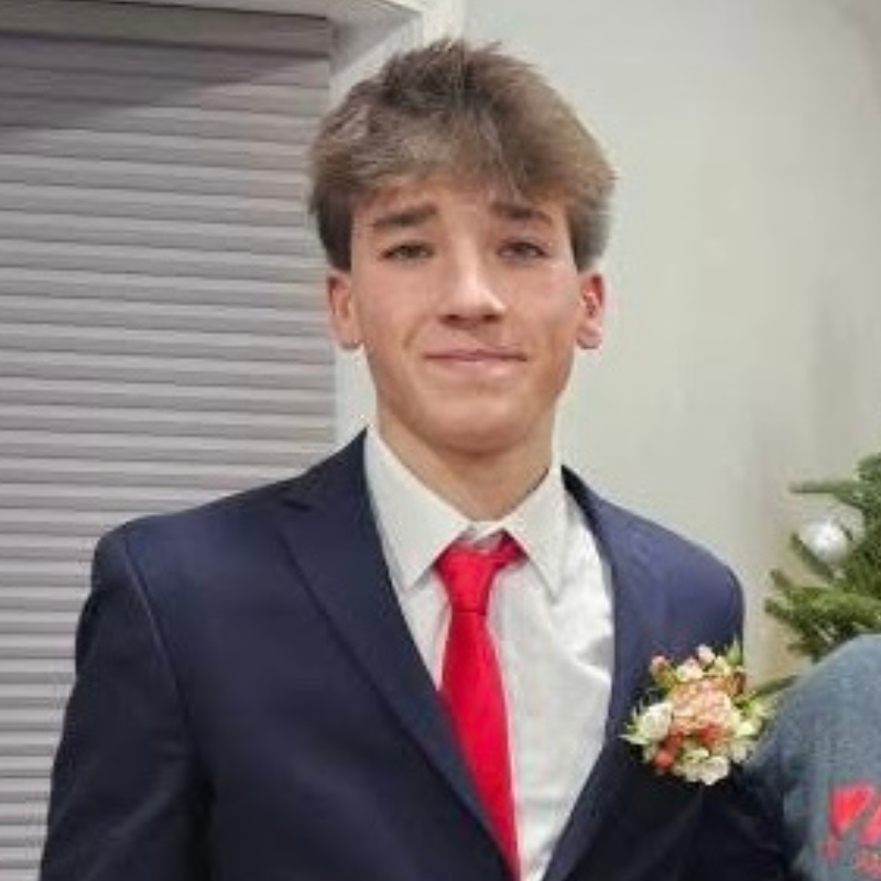 A young man in a dark suit, white shirt, and bright red tie stands indoors. He has short, tousled brown hair and wears a small boutonniere with white and pink flowers on his left lapel.