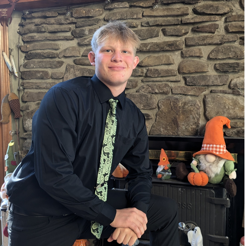 A young man in a black shirt and green patterned tie poses indoors, smiling, with his hand resting on his knee. Behind him is a stone wall and a fireplace decorated with autumn-themed gnome figurines.