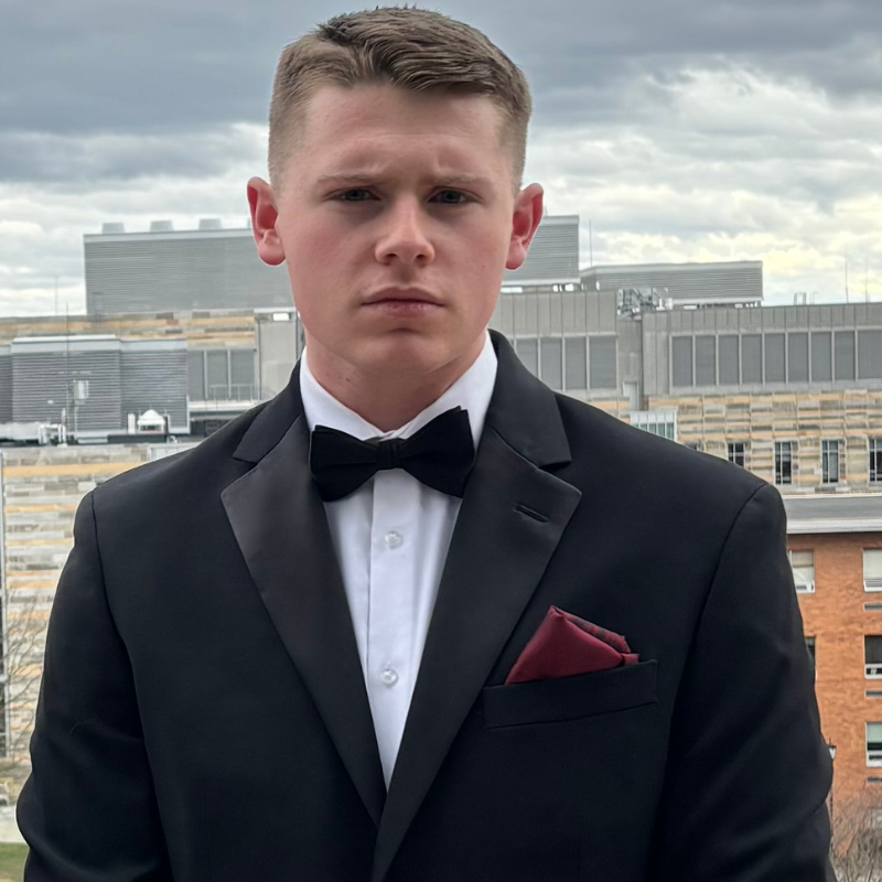 A young man in a black tuxedo with a bow tie and red pocket square stands outdoors against an urban backdrop with cloudy skies and modern buildings in the background.