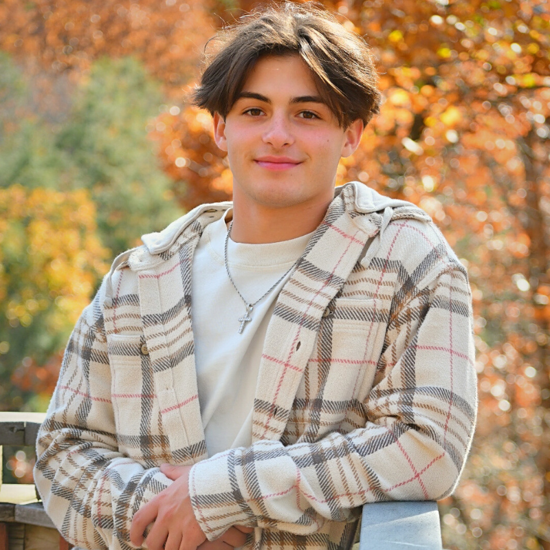 A young man with medium-length brown hair, wearing a plaid jacket and white shirt, leans on a wooden railing outdoors with autumn trees in the background. He is smiling and wearing a cross necklace.