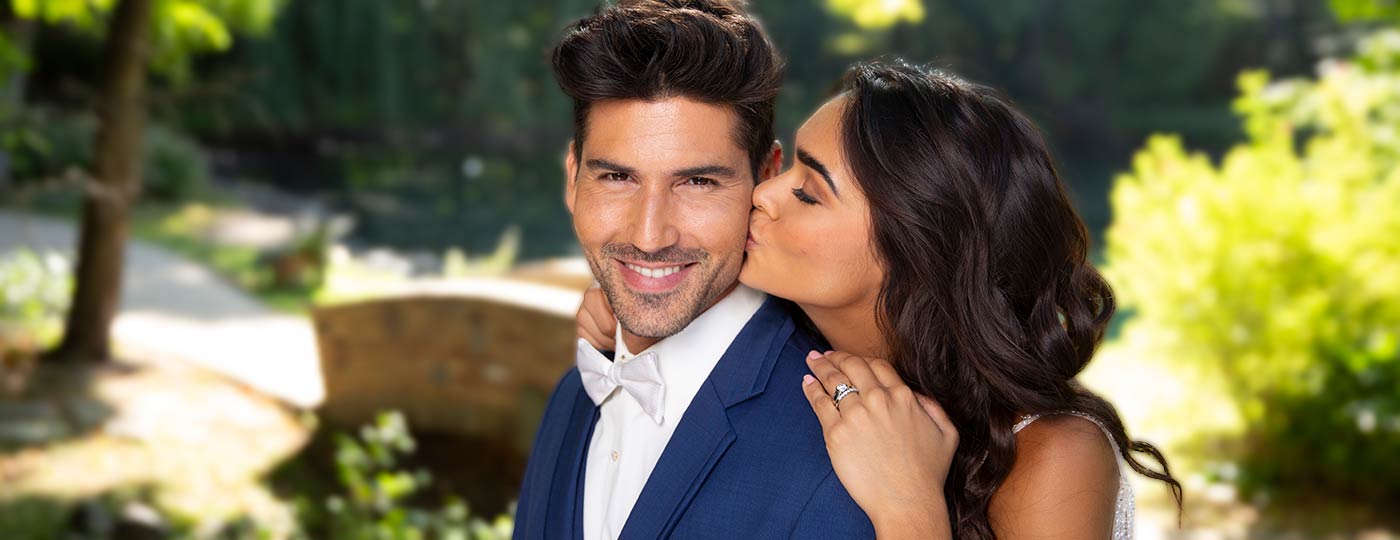 A woman in a white dress kisses a smiling man in a blue suit and bow tie on the cheek. They stand outdoors in a sunny, green park with trees and a blurred stone bridge in the background.