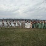 A wedding party poses playfully outdoors in front of water, with groomsmen in beige suits pulling the groom, while bridesmaids in green dresses pull the bride in the opposite direction under a cloudy sky.