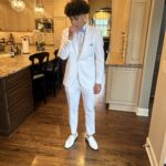 A young man wearing a white suit, white shoes, and a light purple bow tie stands in a modern kitchen with wooden floors and cream-colored cabinets, holding his hand near his face.