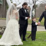A bride in a lace wedding dress stands facing three men in tuxedos and a smiling young boy in a tuxedo, outdoors by a wedding arch decorated with white flowers and tulle. Trees and grass are visible in the background.