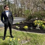 A young man in a black tuxedo, blue bow tie, and sunglasses stands on a driveway near a garden with blooming yellow daffodils and shrubs, under partly cloudy skies with trees in the background.