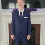 A young man stands indoors in front of a fireplace, wearing a dark blue suit with a white shirt, tie, and pocket square, along with brown dress shoes. He is smiling and posing for the photo.