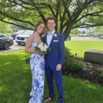A young woman in a white and blue floral dress and a young man in a blue suit with a bow tie stand together smiling, holding a bouquet, on a sunny day in a grassy area with trees and parked cars in the background.