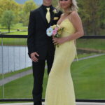 A young couple dressed formally poses outdoors; the young man wears a black suit with a yellow tie, and the young woman wears a fitted yellow gown, holding a bouquet. Green trees and a lake are in the background.
