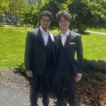 Two young men wearing black tuxedos and bow ties stand side by side, smiling, on a sunny day with green grass and large leafy trees in the background.