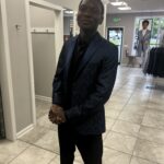 A person stands in a clothing store wearing a dark blue patterned suit jacket and black pants. The person is smiling slightly and has their hands clasped in front. The store interior is bright with racks of suits.