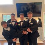 Four young men dressed in black tuxedos and bow ties pose indoors; three of them stand while holding up the fourth horizontally, all smiling in front of a large TV and bright windows.